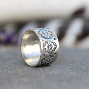 May include: A wide, silver ring with a repeating pattern of starburst designs. The ring is approximately 0.75 inches wide and has a slightly oxidized finish. The ring is displayed on a gray surface with blurred background elements.