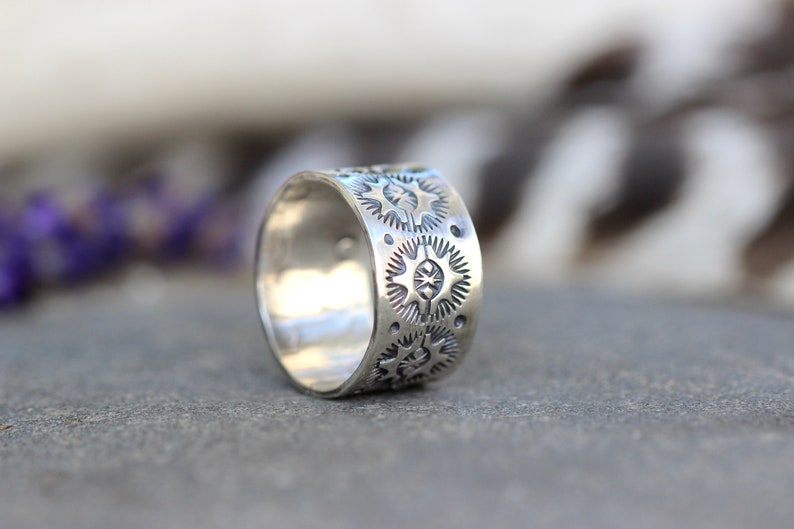 May include: A wide, silver ring with a repeating pattern of starburst designs. The ring is approximately 0.75 inches wide and has a slightly oxidized finish. The ring is displayed on a gray surface with blurred background elements.