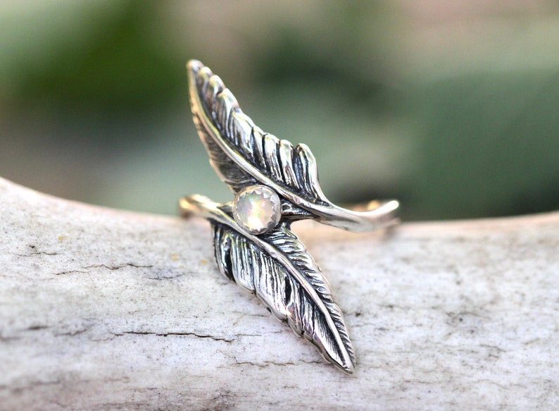 May include: A silver ring with two feather designs and a white opal gemstone in the center.