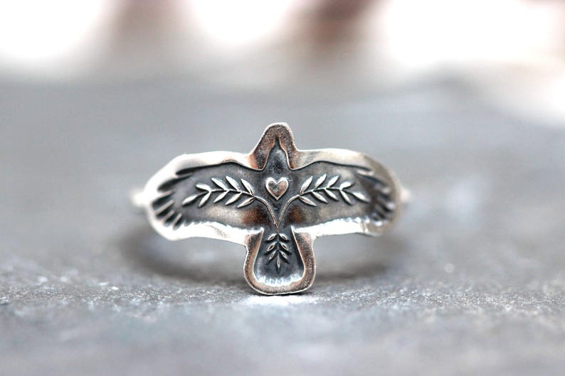 May include: A silver ring featuring a stylized bird design. The bird has outstretched wings with leaf and heart details. The ring is made of a shiny, metallic material.