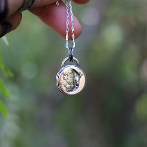 Puede incluir: Un collar de plata con un colgante de luna creciente con una cara y estrellas. El colgante cuelga de una cadena de plata.