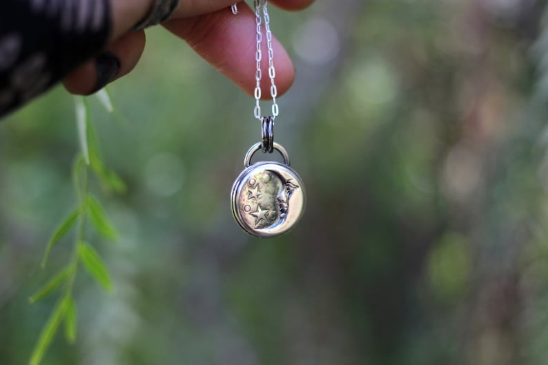 Puede incluir: Un collar de plata con un colgante de luna creciente con una cara y estrellas. El colgante cuelga de una cadena de plata.