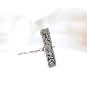 May include: A silver ring with a rectangular band featuring an engraved design of the moon phases.
