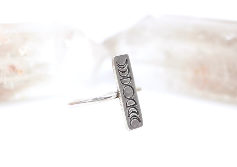 May include: A silver ring with a rectangular band featuring an engraved design of the moon phases.