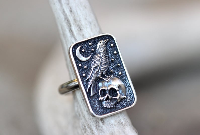 May include: A silver ring with a black rectangular design featuring a raven perched on a skull with a crescent moon and stars in the background.