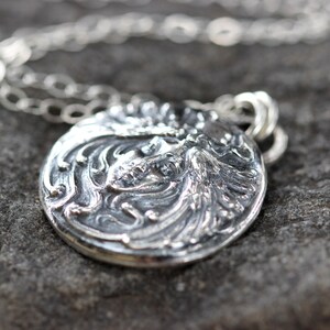 May include: A silver pendant necklace with a detailed, ornate design featuring a woman's face. The pendant is round and has a textured surface.