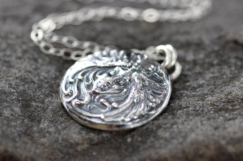 May include: A silver pendant necklace with a detailed, ornate design featuring a woman's face. The pendant is round and has a textured surface.