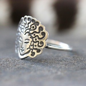 May include: A silver ring with a detailed face design and flowing hair, framed by a decorative border. The ring's face is approximately 2 cm in diameter, with a smooth, polished band. The ring is set against a blurred background.