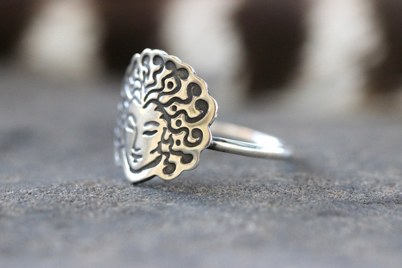 May include: A silver ring with a detailed face design and flowing hair, framed by a decorative border. The ring's face is approximately 2 cm in diameter, with a smooth, polished band. The ring is set against a blurred background.