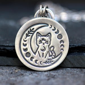 May include: A silver pendant necklace featuring a cat sitting inside a crescent moon with a floral design. The moon is surrounded by smaller crescent moons.