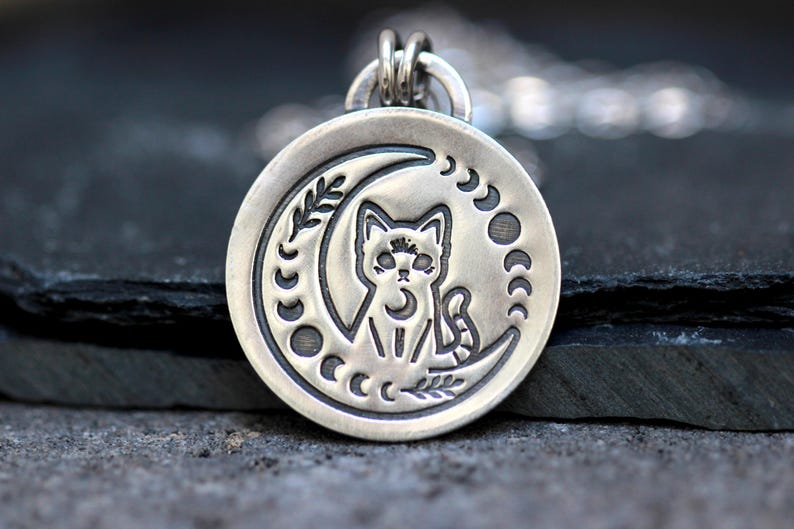 May include: A silver pendant necklace featuring a cat sitting inside a crescent moon with a floral design. The moon is surrounded by smaller crescent moons.