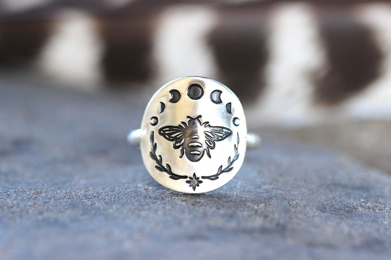 May include: A silver ring with a bee and moon phases design. The bee is centered on the ring and surrounded by five crescent moons. The ring is on a gray background.