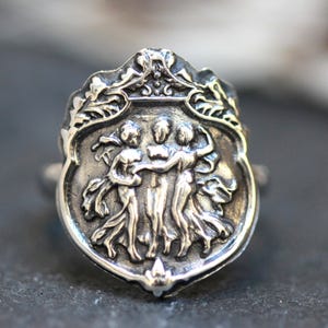 Puede incluir: Un anillo de plata con un dise&ntilde;o detallado de tres figuras que se toman de la mano, rodeado de un borde de hojas.
