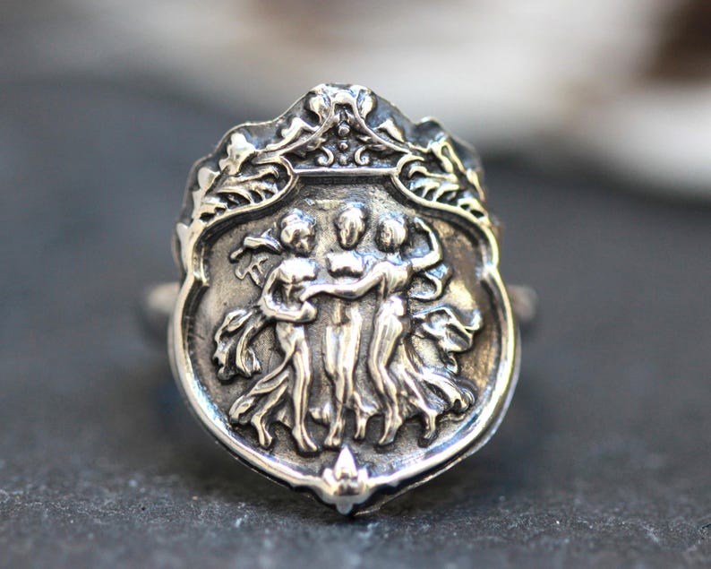 Puede incluir: Un anillo de plata con un dise&ntilde;o detallado de tres figuras que se toman de la mano, rodeado de un borde de hojas.
