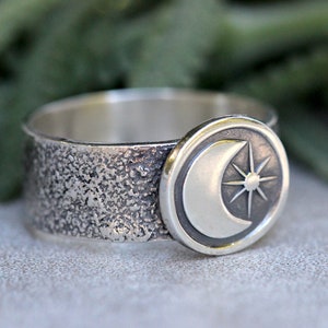 May include: A silver ring with a textured band and a circular design featuring a crescent moon and a star.