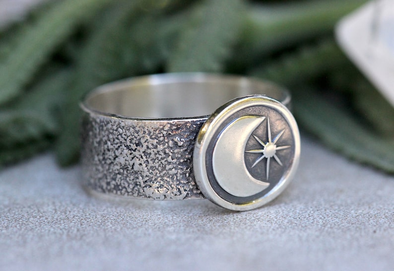 May include: A silver ring with a textured band and a circular design featuring a crescent moon and a star.