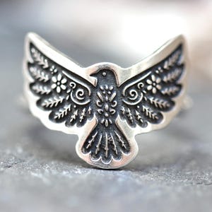 May include: A silver-toned ring featuring a detailed bird design with outstretched wings. The bird's body and wings are adorned with intricate floral and leaf patterns. The ring is set against a textured gray surface.