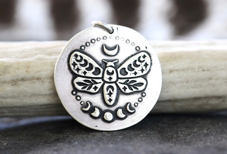May include: A silver pendant featuring a detailed embossed design of a moth with crescent moons and stars on its wings. The pendant is circular and has a textured surface.