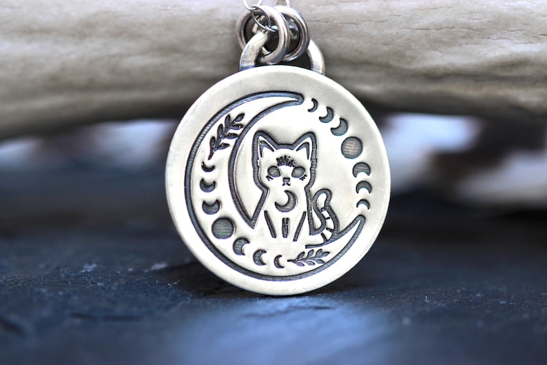 May include: A silver pendant necklace featuring a cat sitting on a crescent moon with a moon phase design. The pendant is round and has a detailed design.