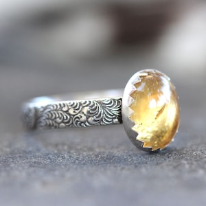 May include: A silver ring with a yellow gemstone set in a scalloped bezel. The band is decorated with a floral pattern.