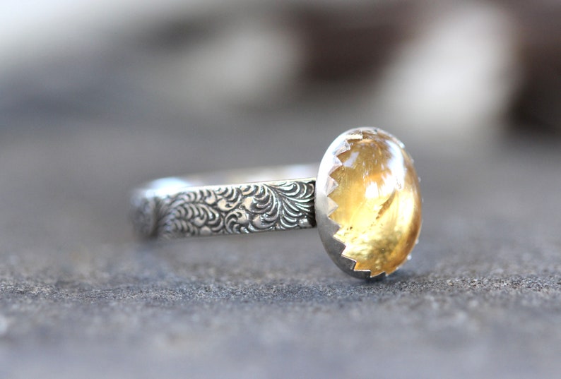 May include: A silver ring with a yellow gemstone set in a scalloped bezel. The band is decorated with a floral pattern.