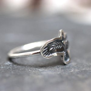 May include: A silver ring with a leaf design. The ring has a thin band and a detailed leaf motif on the face. The ring is shown on a gray surface.