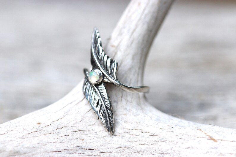 May include: A silver ring with two feather designs and a white opal gemstone in the center.
