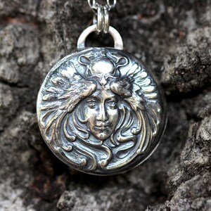 May include: A silver pendant featuring a detailed, embossed design of a woman's face framed by stylised wings. The pendant is round and has a textured surface.