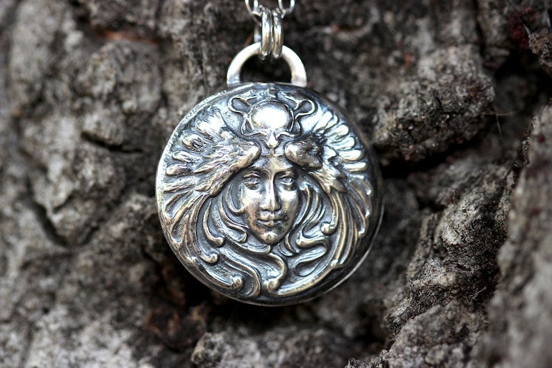 May include: A silver pendant featuring a detailed, embossed design of a woman's face framed by stylised wings. The pendant is round and has a textured surface.