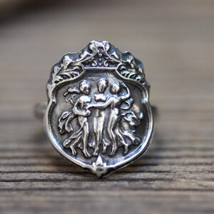 Puede incluir: Un anillo de plata con un dise&ntilde;o detallado de tres figuras que se toman de la mano, ubicado dentro de un marco en forma de escudo con adornos florales ornamentados.
