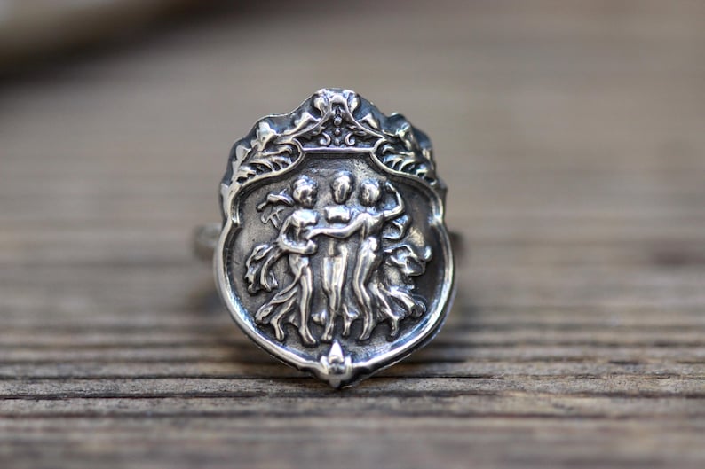 Puede incluir: Un anillo de plata con un dise&ntilde;o detallado de tres figuras que se toman de la mano, ubicado dentro de un marco en forma de escudo con adornos florales ornamentados.