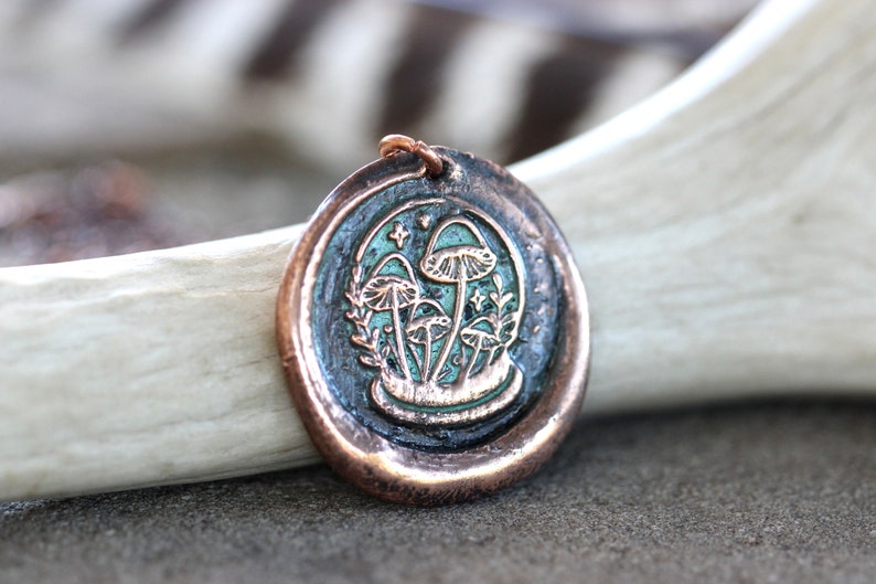 May include: A copper pendant necklace with a green enamel design of mushrooms and leaves. The pendant is round and has a textured surface.