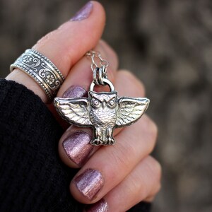 May include: Silver owl pendant necklace with detailed wings. The owl has large, round eyes and a textured surface. The necklace is on a silver chain.