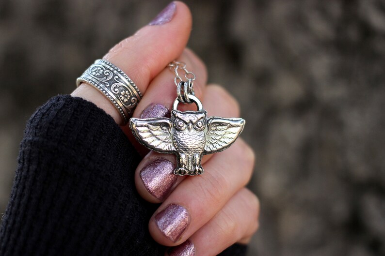May include: Silver owl pendant necklace with detailed wings. The owl has large, round eyes and a textured surface. The necklace is on a silver chain.