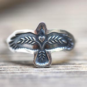 May include: A silver ring featuring a dove design. The dove has outstretched wings, a heart, and olive branch details. The ring is set against a wooden surface, showcasing the intricate details of the jewelry.