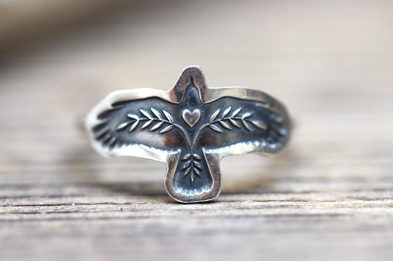 May include: A silver ring featuring a dove design. The dove has outstretched wings, a heart, and olive branch details. The ring is set against a wooden surface, showcasing the intricate details of the jewelry.