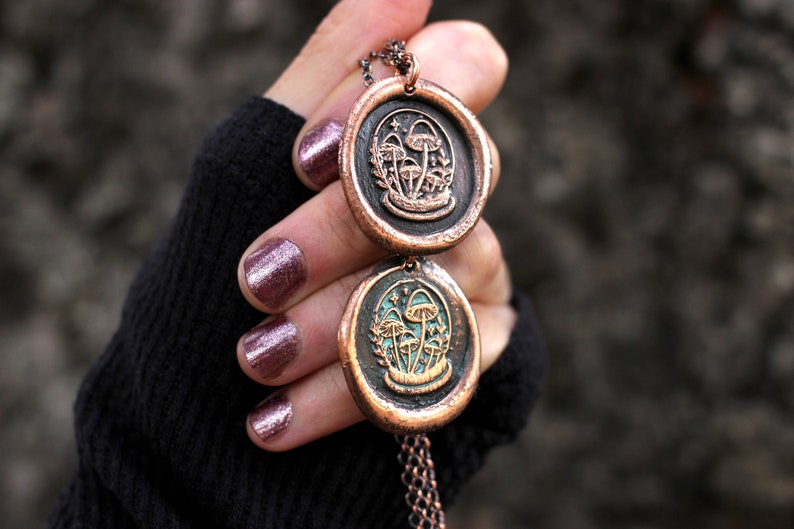 May include: A copper necklace with two round pendants featuring a design of mushrooms and stars. The pendants are connected by a copper chain.