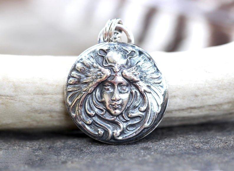 May include: A silver pendant featuring a detailed, ornate design of a woman's face with wings. The pendant is round and has a textured surface.
