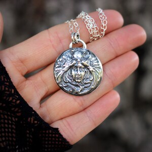 May include: A silver pendant necklace featuring a detailed, ornate design of a woman's face surrounded by stylised leaves and flowers.