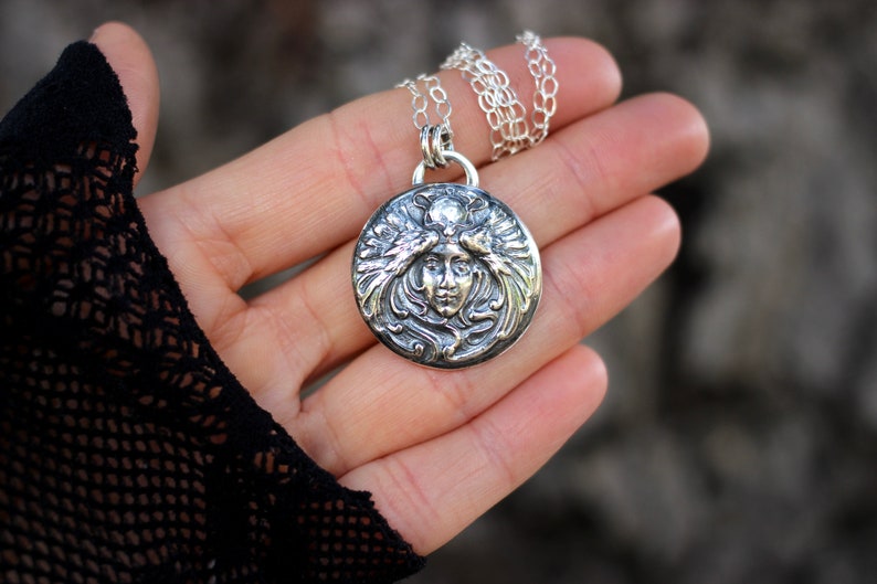 May include: A silver pendant necklace featuring a detailed, ornate design of a woman's face surrounded by stylised leaves and flowers.