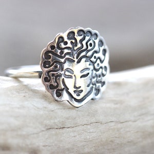 May include: A silver ring with a detailed face design and flowing hair. The face has closed eyes and a serene expression. The ring is made of a shiny metal and is set against a neutral background.