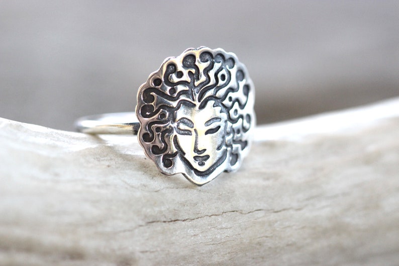 May include: A silver ring with a detailed face design and flowing hair. The face has closed eyes and a serene expression. The ring is made of a shiny metal and is set against a neutral background.