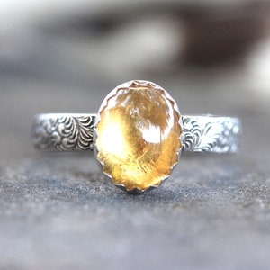 May include: A silver ring with an oval-shaped, yellow citrine gemstone set in a scalloped bezel. The band is decorated with a floral design.