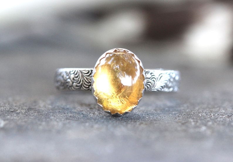 May include: A silver ring with an oval-shaped, yellow citrine gemstone set in a scalloped bezel. The band is decorated with a floral design.
