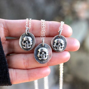 May include: Three silver-toned skull pendants on delicate chains. Each pendant features a detailed skull within an oval frame. The necklaces are displayed against a blurred background, highlighting the jewelry's intricate design. The chains are of varying lengths.