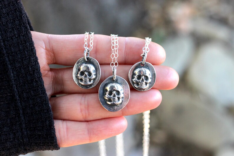 May include: Three silver-toned skull pendants on delicate chains. Each pendant features a detailed skull within an oval frame. The necklaces are displayed against a blurred background, highlighting the jewelry's intricate design. The chains are of varying lengths.