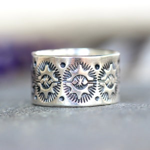 May include: A wide, silver ring with a detailed pattern of star-like shapes and small dots. The ring has a slightly oxidized finish, giving it a vintage or antique appearance. The design is repeated around the entire band.