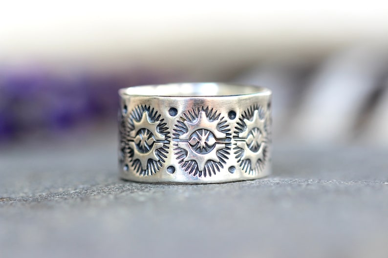 May include: A wide, silver ring with a detailed pattern of star-like shapes and small dots. The ring has a slightly oxidized finish, giving it a vintage or antique appearance. The design is repeated around the entire band.