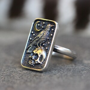 May include: A silver ring with a rectangular design featuring a detailed depiction of a raven perched on a skull with a crescent moon and stars in the background.