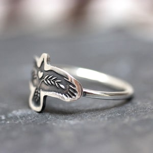 May include: A silver ring featuring a detailed bird design. The bird's head is visible, with a heart and leaf details. The ring is set against a gray background, showcasing the intricate craftsmanship of the jewelry.